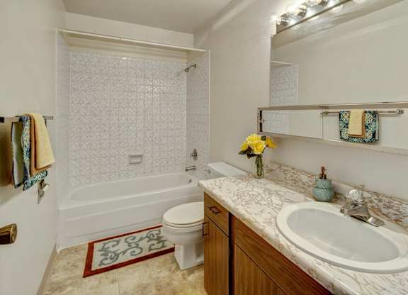 A clean bathroom at Arctic Sun Apartments featuring a bathtub and shower combo with tiled walls, a toilet, and a long marble-style countertop with a sink. A wide mirror with vanity lighting brightens the space, and towel racks and decor add color and warmth.