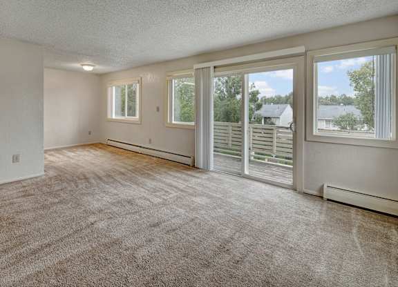 A bright living space at Arctic Sun Apartments with plush beige carpet, neutral walls, and large windows. A sliding glass door opens to a private balcony with neighborhood views, bringing in abundant natural light. The open layout offers flexible room for seating and dining arrangements.