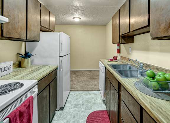 A galley kitchen at Arctic Sun Apartments with wood cabinets, long laminate countertops, a double sink, and a stovetop. A refrigerator and dishwasher offer added convenience. The space opens into a dining nook with carpeted flooring, creating a functional layout for cooking and meals.