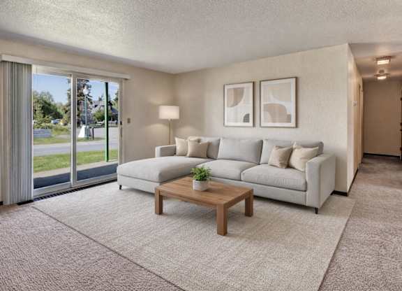 A bright living room at Bay Arms Apartments with a large sectional sofa, a wooden coffee table, and neutral décor. A sliding glass door opens to a grassy outdoor area and lets in natural light. Soft carpet and a hallway leading to the bedrooms create a comfortable open layout.