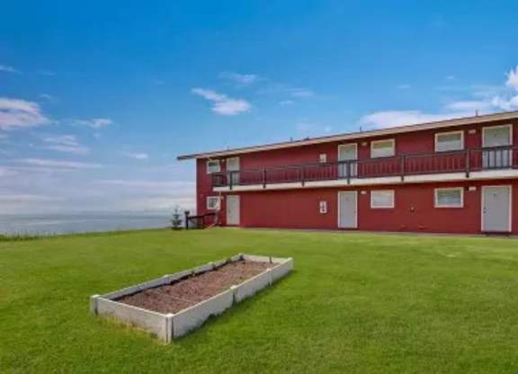 A scenic exterior view of Bay Arms Apartments featuring a two-story red building with balconies and white trim. A wide grassy lawn stretches across the property with a raised planting bed in the center. The ocean sits just beyond the grounds, offering peaceful waterfront views.