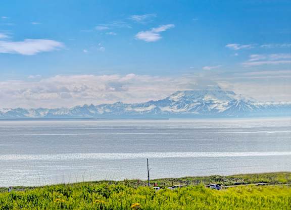 A stunning waterfront view at Bay Arms Apartments overlooking calm ocean water and a sweeping mountain range in the distance. Bright blue skies and green shoreline vegetation create a peaceful, scenic backdrop just steps from the community.