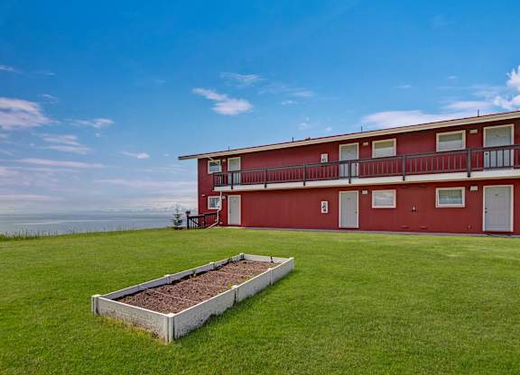 A scenic exterior view of Bay Arms Apartments featuring a two-story red building with balconies and white trim. A wide grassy lawn stretches across the property with a raised planting bed in the center. The ocean sits just beyond the grounds, offering peaceful waterfront views.