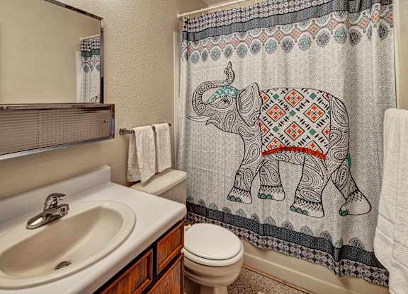 A cozy bathroom at Bay Arms Apartments with a vanity and sink, a wide mirror, and a shower and bathtub combo. A patterned curtain with an elephant design adds color and personality. The room includes a toilet, towel rack, and neutral walls, creating a clean and welcoming space.