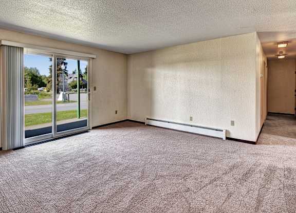 A spacious living room at Bay Arms Apartments with plush carpet and neutral walls. A large sliding glass door opens to a grassy outdoor area and fills the space with natural light. A hallway leads to the bedrooms and bathroom, offering an open and functional layout.
