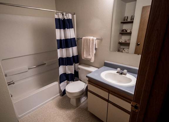 A tidy Birchwood bathroom featuring a white bathtub with a bold navy and white striped shower curtain. A compact vanity with a blue countertop and single sink sits beneath a wide mirror. Soft towels hang on the wall, and open shelving stocked with neatly rolled linens adds simple, useful storage.