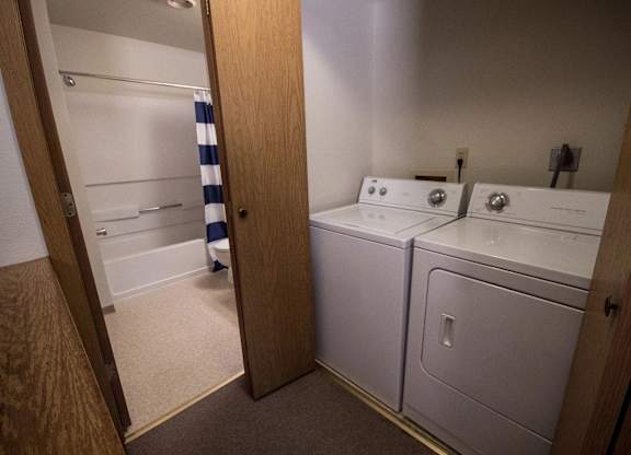 A functional Birchwood laundry nook featuring a white top-load washer and dryer tucked behind sliding wooden doors, with the bathroom just steps away. The adjoining bath includes a tub with a bold navy and white striped shower curtain, creating a practical and organized household layout.