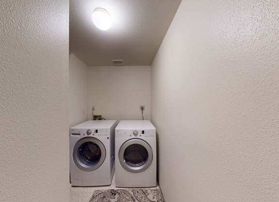 A simple Birchwood laundry room with a front-loading washer and dryer placed side by side at the end of a narrow nook. Soft neutral walls and overhead lighting brighten the compact space, while a patterned rug adds a touch of comfort beneath the appliances.