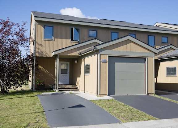 A well-kept Birchwood townhome exterior featuring a tan two-story façade with gray trim, a single-car garage, and a small covered entryway with a white front door. The freshly paved driveway and green lawn frame the home, while a tree and bright summer skies add curb appeal and a welcoming neighborhood feel.