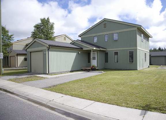 A bright Birchwood townhome with a sage-green exterior, attached single-car garage, and covered entryway sits on a wide open lawn under partly cloudy skies. Multiple vertical windows line the façade, and a small flower planter frames the front steps, giving the home a tidy and inviting curb appeal.
