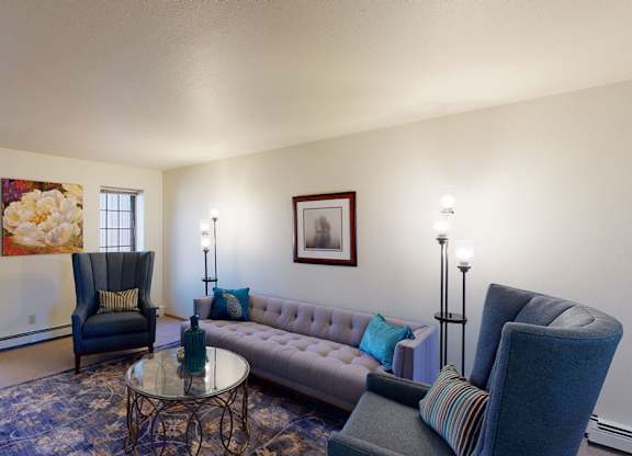 A bright living room at Birchwood Apartments with a tufted sofa, two upholstered high-back chairs, and a round glass coffee table on a patterned area rug. Wall art, accent pillows, and floor lamps add color and warmth, while two windows bring in natural light from outside.