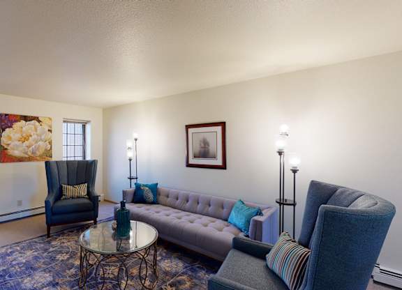A bright living room at Birchwood Apartments with a tufted sofa, two upholstered high-back chairs, and a round glass coffee table on a patterned area rug. Wall art, accent pillows, and floor lamps add color and warmth, while two windows bring in natural light from outside.