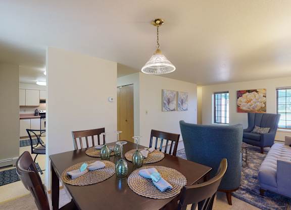 Dining area at Birchwood Apartments featuring a dark wood table set for four with woven placemats and folded napkins, opening into a cozy living room with upholstered seating, floral artwork, and large windows letting in natural light.