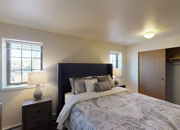 A bright Birchwood bedroom featuring a dark upholstered headboard with paisley bedding and layered pillows centered against a neutral wall. Two matching lamps on wooden nightstands sit beside the bed beneath twin windows. The room includes soft carpet and a wide closet with sliding wooden doors partially open.