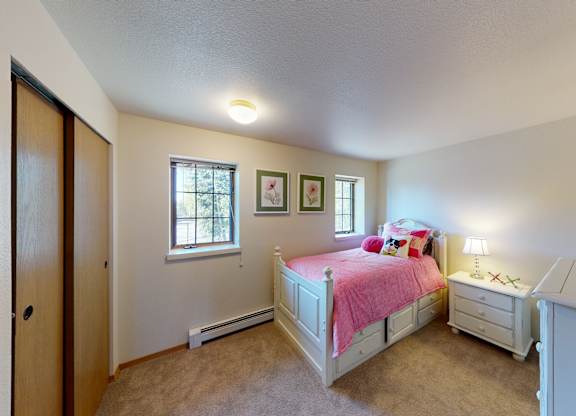 A cheerful Birchwood bedroom with soft beige walls and carpet, featuring a white twin bed with storage drawers and bright pink bedding topped with playful accent pillows. Two windows let in natural light above floral wall art. A matching nightstand with a lamp and a coordinating dresser complete the cozy space beside a wide closet with sliding wooden doors.