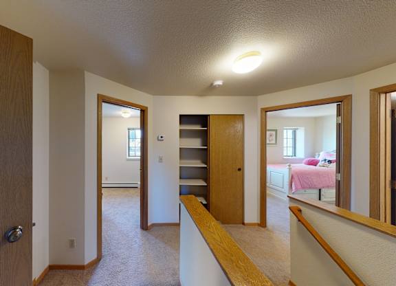 A clean Birchwood hallway with soft carpet and warm wood trim leading to multiple bedrooms. The landing features a built-in shelving niche with sliding closet doors and openings to rooms lit by natural light. A wooden handrail lines the staircase edge, and glimpses of neutral walls and simple finishes create a tidy, open transition space.