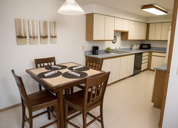 Dining space at Birchwood Apartments featuring a wooden table set for four with rolled napkins, opening to a spacious kitchen with light cabinetry, long countertops, and modern appliances.