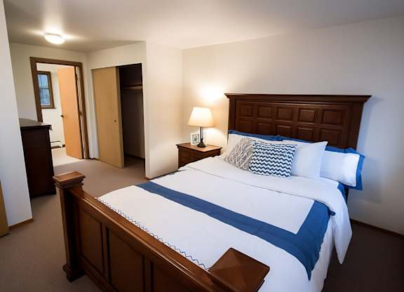 A cozy Birchwood bedroom featuring a wooden bed frame with paneled headboard, dressed in crisp white bedding accented with blue trim and patterned throw pillows. A warm lamp sits on the matching nightstand beside a framed photo. The room includes soft carpet, an open closet with sliding doors, and a doorway leading to the hall.