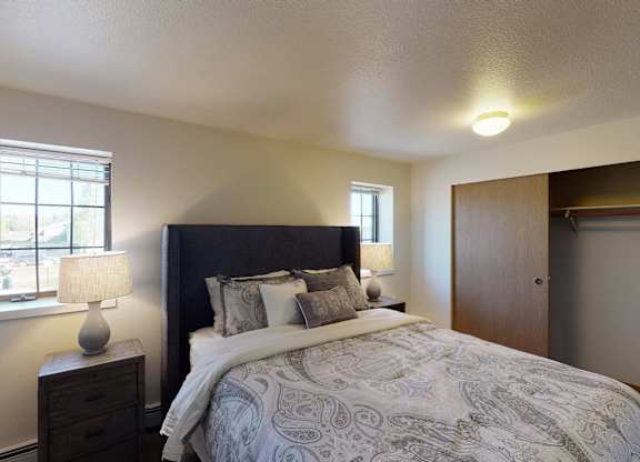 A bright Birchwood bedroom featuring a dark upholstered headboard with paisley bedding and layered pillows centered against a neutral wall. Two matching lamps on wooden nightstands sit beside the bed beneath twin windows. The room includes soft carpet and a wide closet with sliding wooden doors partially open.