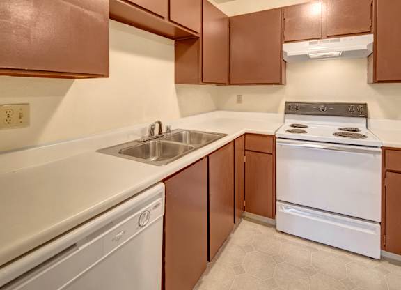 A cozy Brighton Woods kitchen with warm brown cabinets, white countertops, and matching white appliances, including a dishwasher and electric stove. The counters wrap around in an L-shape, providing prep space beside the double stainless-steel sink, with soft neutral walls and flooring framing the cozy, functional layout.