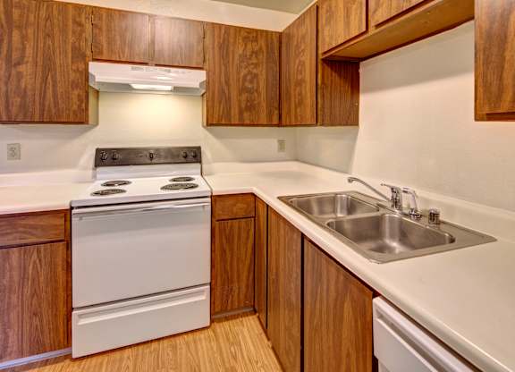 A warm Brighton Woods kitchen featuring wood-grain cabinets, white countertops, and matching white appliances. The electric stove sits beneath a vent hood, while a double stainless-steel sink lines the L-shaped counter. Soft neutral walls and light flooring make the space bright and functional for daily cooking and cleanup.