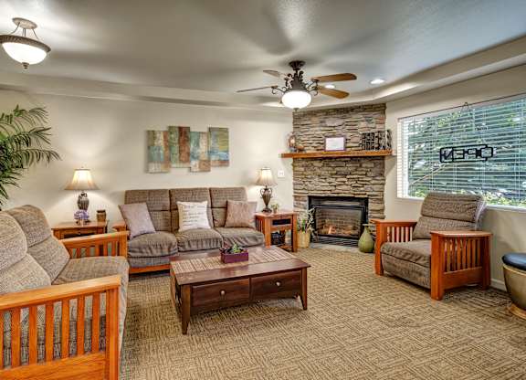 A warm community lounge here at Castle featuring plush seating with wood-framed sofas and armchairs arranged around a wooden coffee table. A stone fireplace with a mantel adds a cozy focal point, while lamps, wall art, and large windows create a welcoming space for residents to relax and gather.