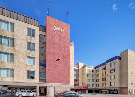 A bright exterior view here at Castle showing the multi-story beige and red building beneath a clear blue sky. Large windows line the façade, with the bold red central tower displaying the Castle name. Doorways, parking areas, and cars are visible at ground level, creating a welcoming first impression of the community.