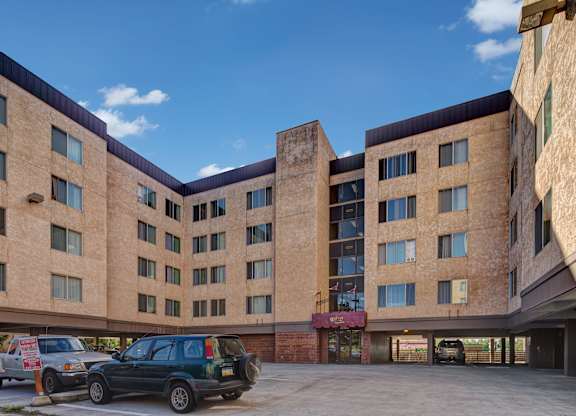 A wide exterior view here at Castle featuring a multi-story beige building with rows of windows and covered parking below. The entrance sits beneath a maroon awning at the center, with parked cars in the foreground and a bright blue sky with soft clouds above the community.