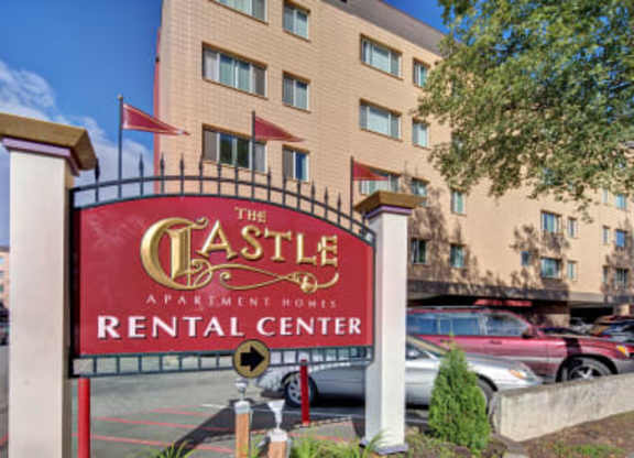 A welcoming exterior view here at Castle showcasing the bold red rental center sign framed by white pillars and decorative fencing in the foreground. Behind it rises the community’s beige multi-story building with large windows, fluttering red flags, and resident parking beneath a sunny blue sky.