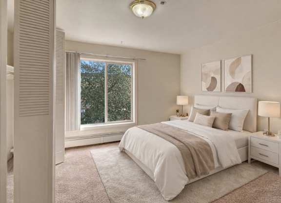 A serene bedroom here at Castle showcases a neatly made bed with white bedding, soft tan throws, and coordinating pillows set against two modern abstract prints. Plush carpet runs wall to wall while large windows bring in natural light and showcase leafy treetop views beside warm bedside lighting on matching nightstands.