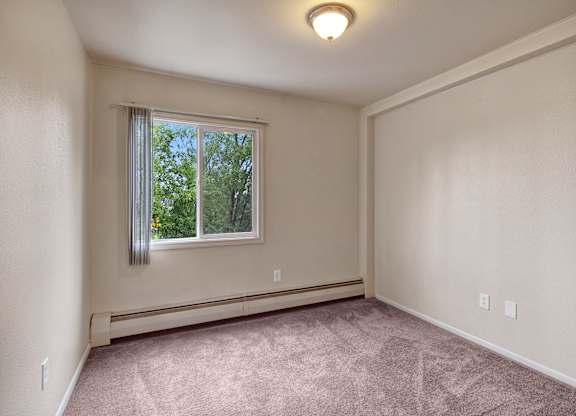 A cozy bedroom here at Castle features soft neutral walls and plush carpeting that creates a warm and inviting feel. A large window brings in natural light and frames lush green treetops outside, while simple baseboard heating and an uncluttered layout offer a versatile space ready for personal furniture and décor.