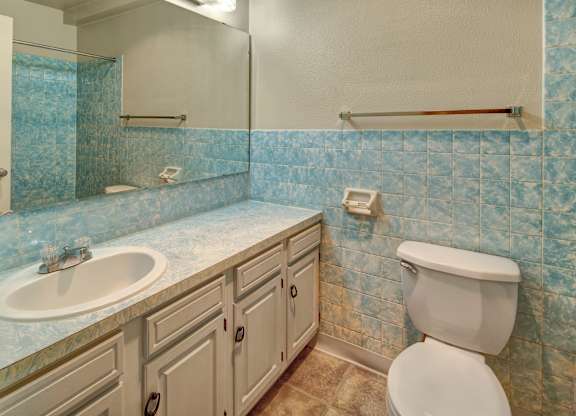 A bright bathroom here at Castle featuring light blue marbled tile walls, a long countertop with matching blue pattern, a white sink and vanity with storage, and a standard toilet. A wide mirror spans the length of the vanity, reflecting the light and giving the room an open, airy feel.