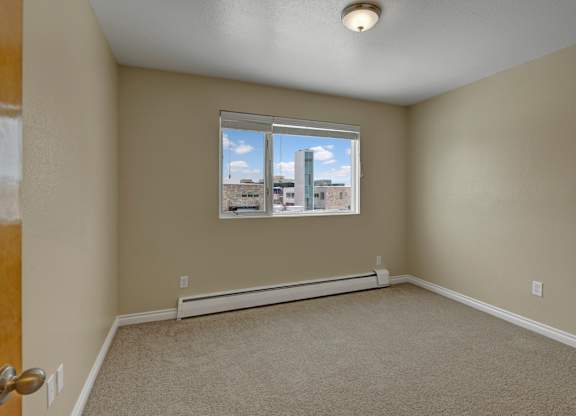 A clean bedroom here at Century Plaza featuring soft beige carpeting and warm neutral walls. A wide window fills the room with natural daylight and provides a view of nearby buildings and the bright blue sky outside. The simple layout offers flexibility for furniture placement and décor.