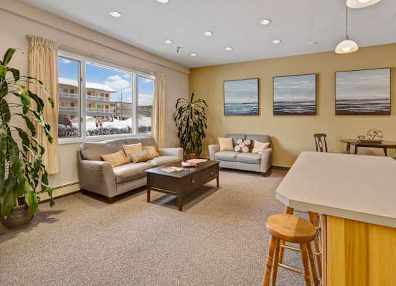 A bright community lounge here at Century Plaza featuring two cozy loveseats with soft throw pillows, a dark wood coffee table, and large windows bringing in natural light. Three coastal art pieces line the yellow accent wall, with plants, bar seating, and a small dining table completing the inviting space.