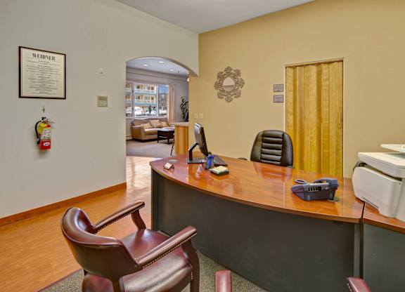 A welcoming leasing office lobby here at Century Plaza featuring a curved wood desk with a computer, phone, and office chair, plus guest seating, warm neutral walls, and an arched opening leading to a cozy community lounge with sofas and large windows.