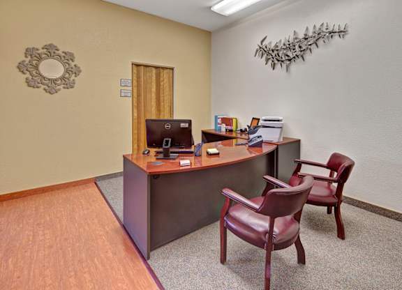 A clean and professional leasing office space here at Century Plaza featuring a curved wood desk with a computer and office essentials, two leather guest chairs, neutral walls with decorative accents, and bright overhead lighting.