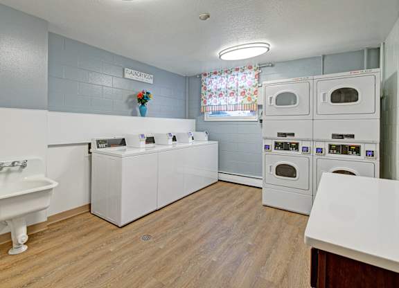 A bright on-site laundry room here at Century Plaza featuring multiple top-loading washers, stacked dryers, a deep utility sink, wood-look flooring, and a window with colorful curtains for natural light.