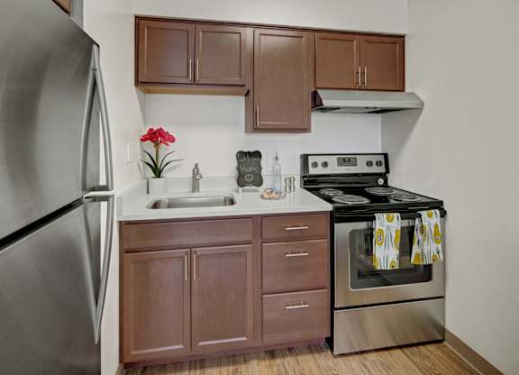 A compact modern kitchen here at Century Plaza with sleek stainless-steel appliances, rich wood cabinets, and a bright white countertop. A stainless sink, electric range, and overhead vent provide everyday convenience, while simple décor and wood-style flooring create a clean and cheerful cooking space.