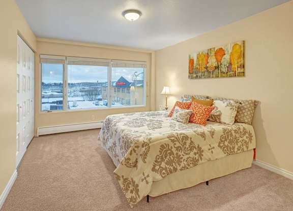 A spacious bedroom here at Century Plaza featuring a large window with sweeping neighborhood views, soft neutral carpeting, and warm cream walls. A queen bed with patterned bedding and colorful accent pillows sits beneath wall art, creating an inviting and comfortable retreat filled with natural light.