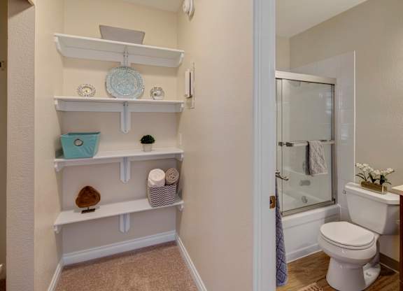 A stylish nook and bathroom space here at Century Plaza featuring open white shelving neatly decorated with plates, towels, and woven baskets to maximize storage. Off to the right, the bathroom includes a glass-door shower and tub, a white toilet, and warm natural lighting for a clean, organized feel.