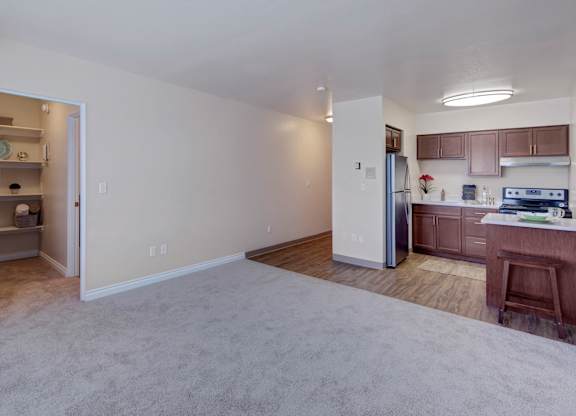A spacious living area here at Century Plaza showcasing an open layout with plush carpet flowing into warm wood-style flooring in the kitchen. The kitchen features rich espresso cabinets, stainless-steel appliances, and a small breakfast bar, while a built-in shelving nook off to the side adds convenient storage and display space.