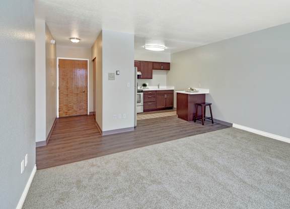A spacious living area here at Century Plaza featuring a blend of soft carpet and rich wood-style flooring that transitions into the kitchen. The open layout includes a breakfast bar with stool seating, warm wood cabinetry, and modern lighting, while the front entry hallway adds convenience and flow to the space.