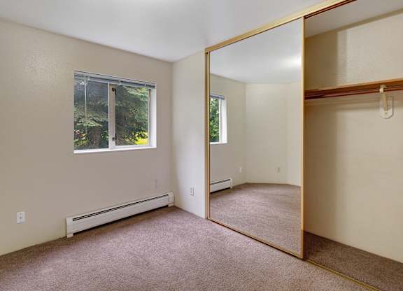 A bright empty bedroom here at Chugach South with beige walls, carpet flooring, large mirrored closet doors, and a window looking out to trees.