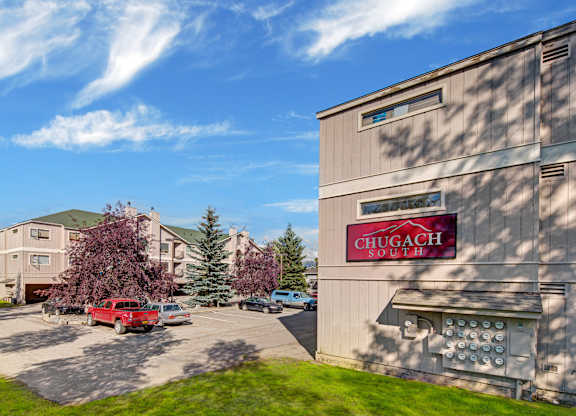 A sunny exterior view of Chugach South featuring tan apartment buildings with a red property sign, parking lot with several vehicles, green lawn, and tall trees under a blue sky with wispy clouds.