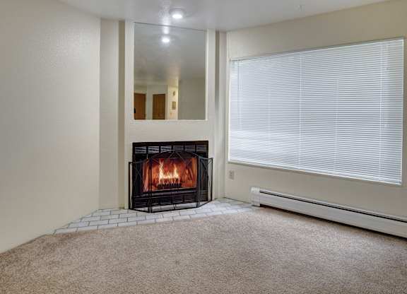 A bright living room here at Chugach South featuring beige carpeting, a lit wood-burning fireplace with a decorative screen and mirror above it, and a large window with white blinds.