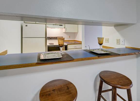 A cozy kitchen peek here at Chugach South showing a raised breakfast bar with two wooden stools, place settings with glasses, and a view into the kitchen with white appliances and cabinets.