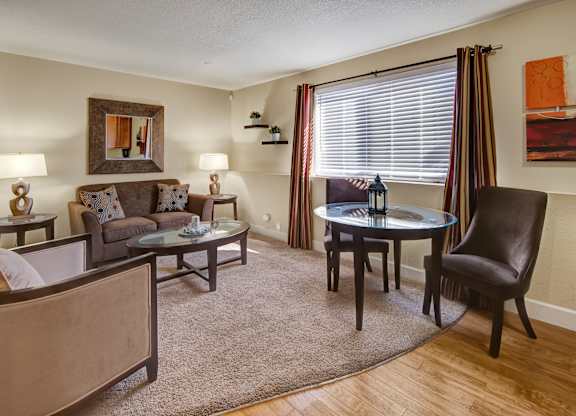 A warm furnished living room here at City View featuring a brown sofa with patterned pillows, matching chairs, glass top coffee and dining tables, floor and table lamps, wall art, and a large window with striped curtains letting in natural light.