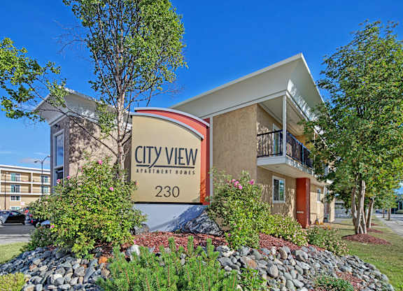 A welcoming front view of City View Apartment Homes featuring the large entrance sign surrounded by colorful landscaping with rocks, shrubs, and blooming flowers. Modern tan buildings with balconies rise behind it, and a bright blue sky adds a cheerful backdrop to this well-kept community.