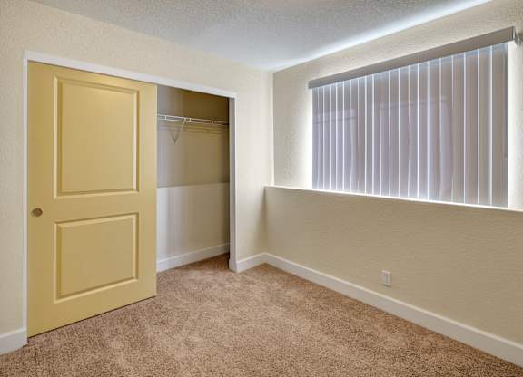 A bright bedroom here at City View featuring soft tan carpeting, neutral walls, and a large window with vertical blinds that filters natural sunlight into the space. A wide closet with sliding doors provides easy storage and an open layout offers flexibility for bedroom furniture or a cozy work-from-home setup.