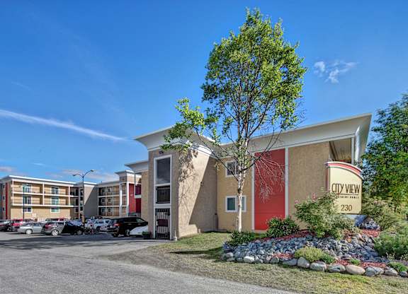 A sunny exterior view of City View Apartment Homes showcasing modern multi-story buildings with bold red and tan accents, balconies, and well-kept landscaping framed by rocks, shrubs, and trees. A clear blue sky, parked cars, and the welcoming entrance sign create a bright and inviting first impression of the community.